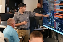 people sitting in chairs and listen to ta person standing, pointing at a  large computer screen.