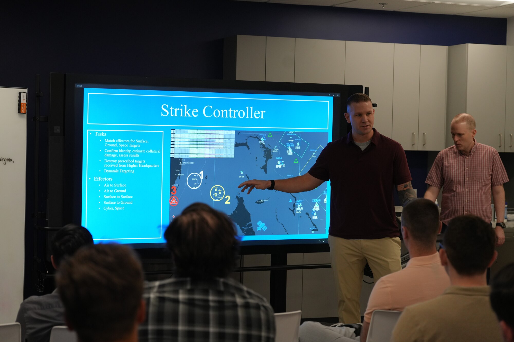 civilians sitting in chairs and standing listen to the leader standing, there is large computer screen mounted on the wall behind the people and people sitting working on computers in the room