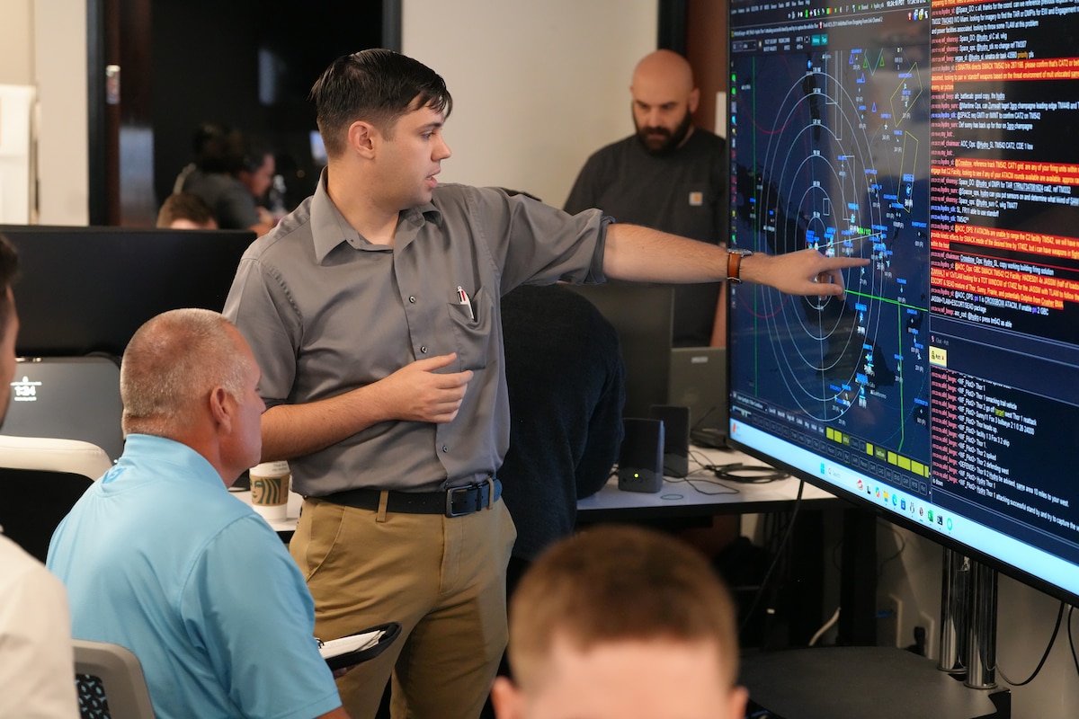 people sitting in chairs and listen to ta person standing, pointing at a  large computer screen.