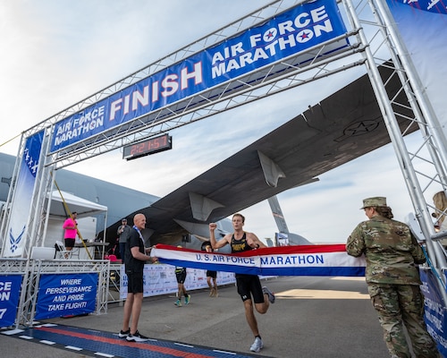 A runner prepares to break a red, white, and blue finish tape with a person on either side. The banner reads U.S. Air Force Marathon.