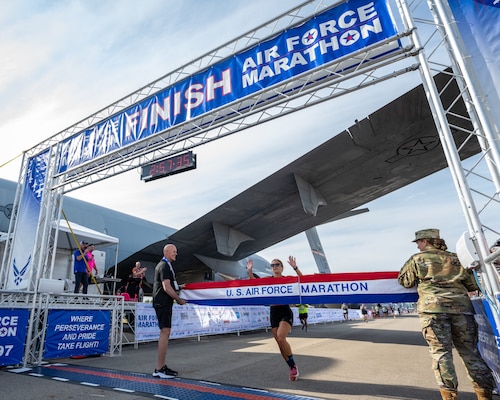 A runner prepares to break a red, white, and blue finish tape with a person on either side. The banner reads U.S. Air Force Marathon.
