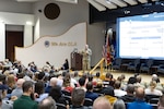 A person in a military uniform stands on stage explaining concepts to a packed auditorium.