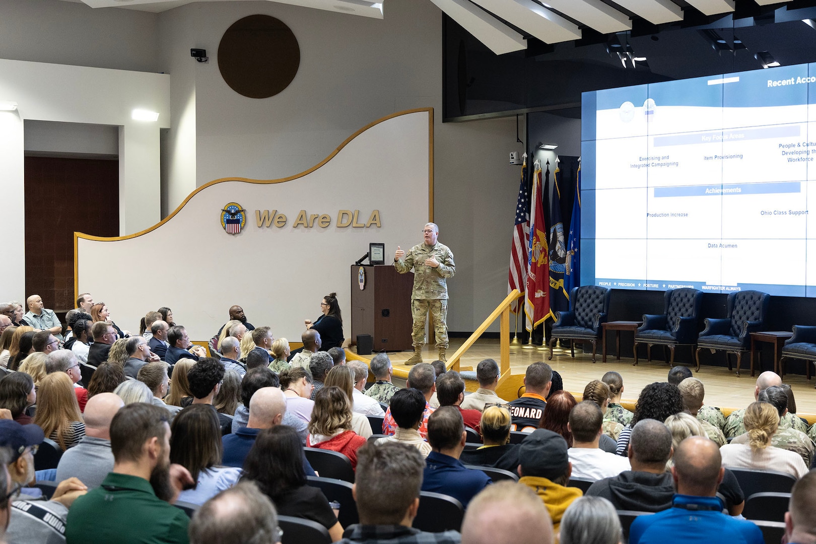 A person in a military uniform stands on stage explaining concepts to a packed auditorium.