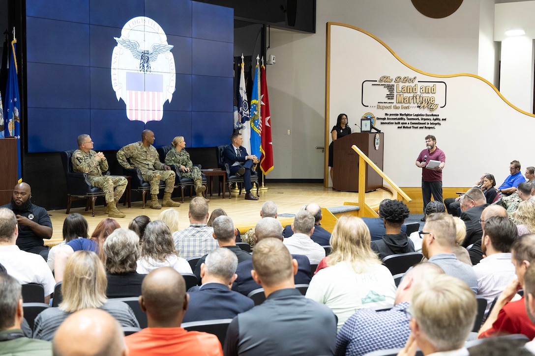Three people in military uniforms and one person in a suit sit on stage and listen to a question from the audience. A person in a black dress stands at the podium in the background. .
