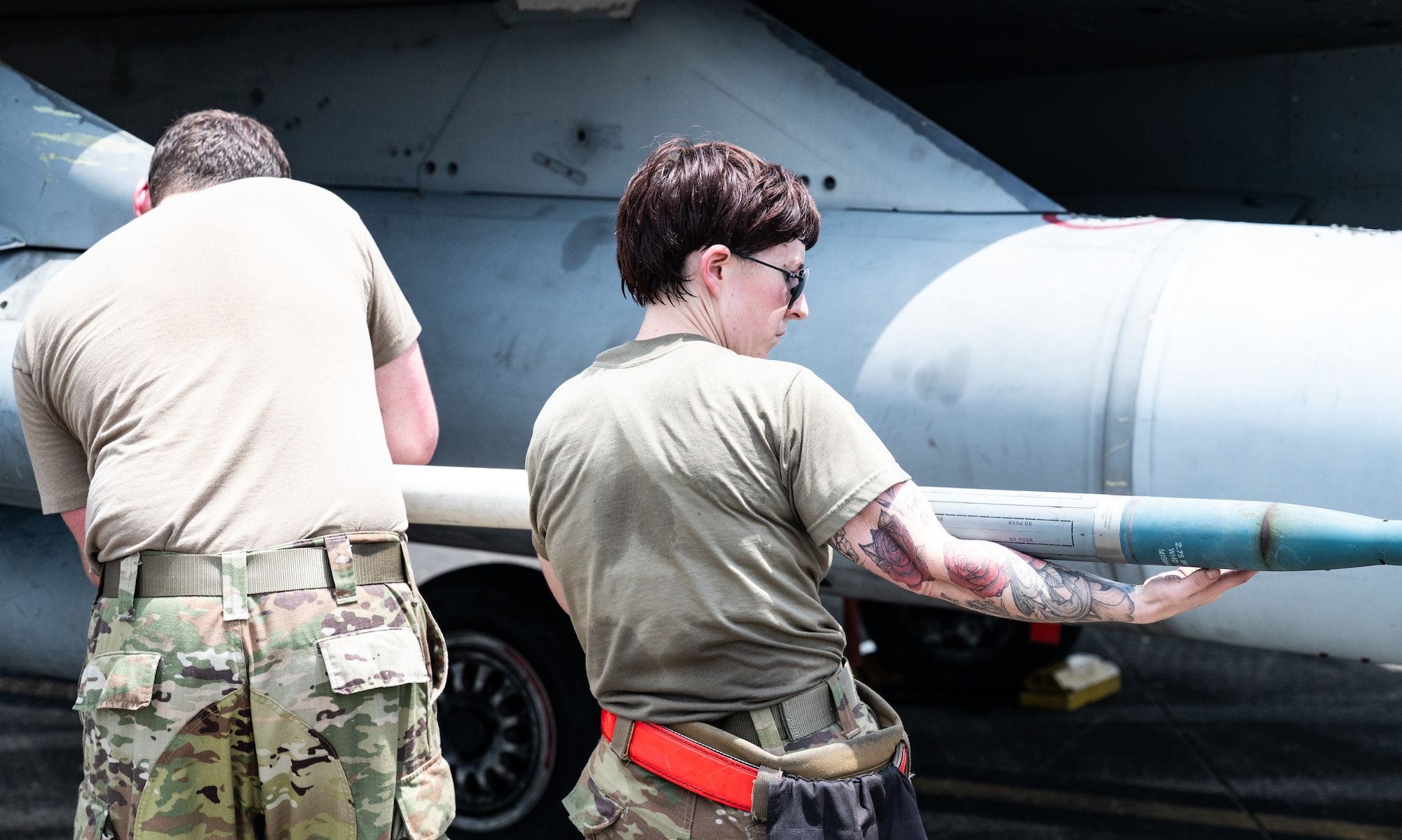 Two maintainers, one male, one female, face away from the camera, loading a rocket into a launcher on a jet.