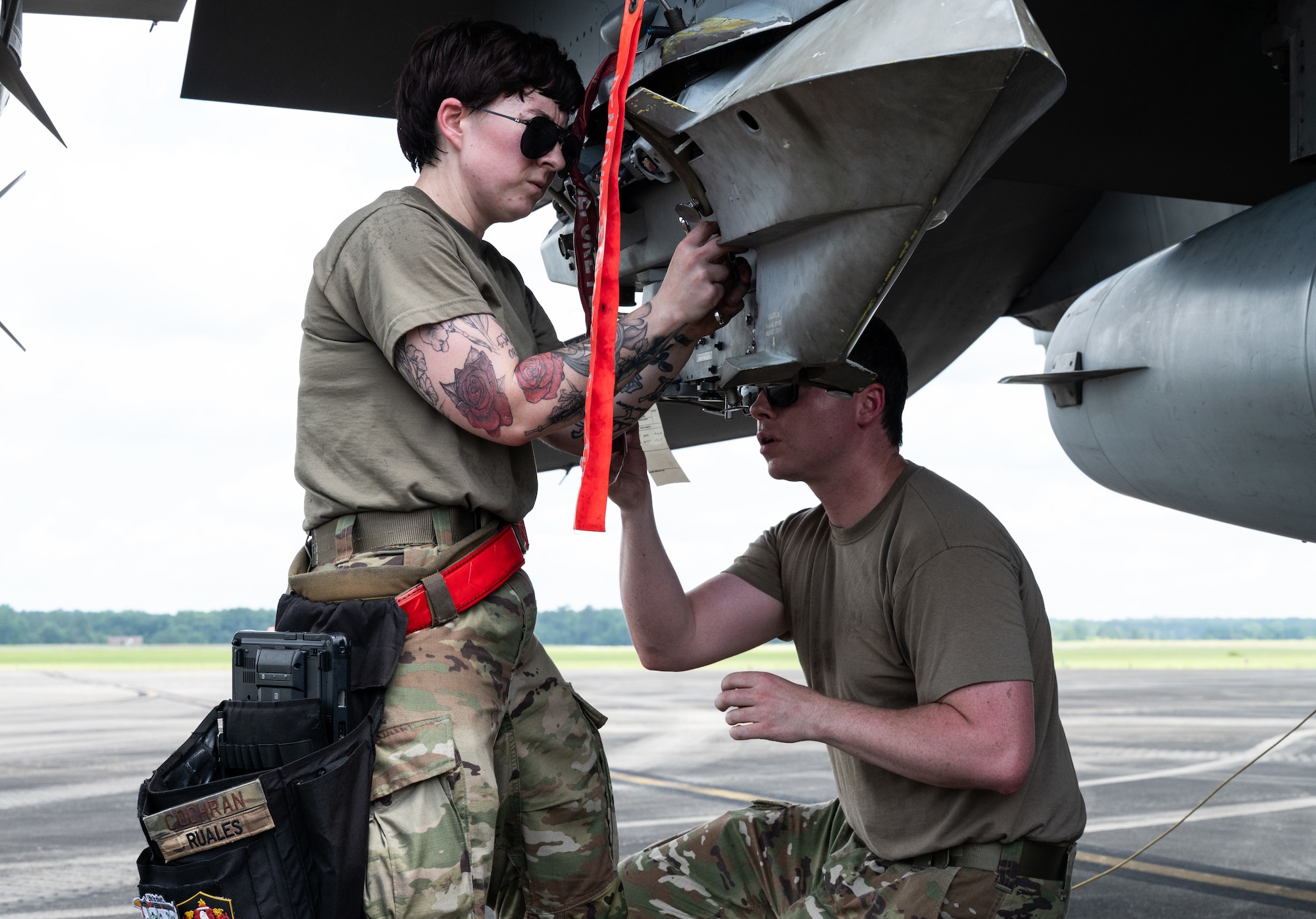 A female maintainer turns a wrench on a rocket pod attached to a jet while a male maintainer crouches on the other side, ensuring the munition holds steady.