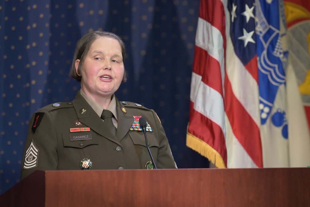 An Army Command Sergeant Major speaks at a podium with military flags behind her.