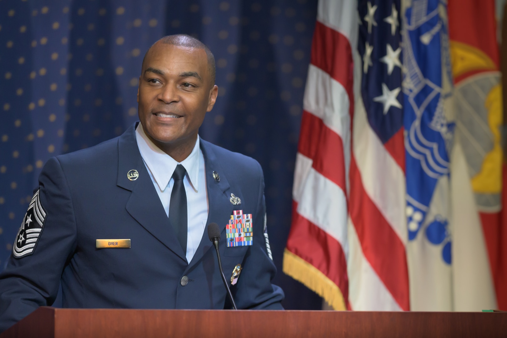 An Air Force Command Chief Master Sergeant speaks at a podium with military flags behind him.