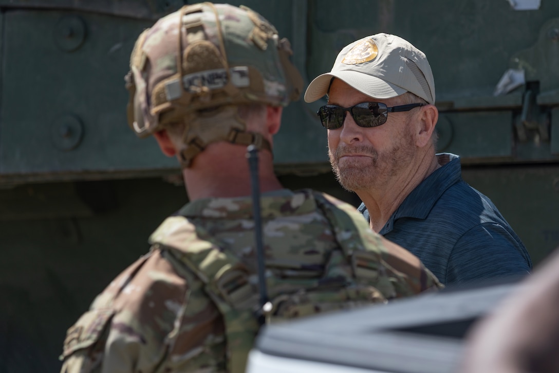 A person in sunglasses and a baseball cap looks at a soldier whose back is turned during the day.