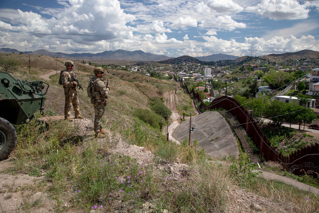 Two soldiers stand on a grassy hill and look over a fence at a city under a blue sky dotted with clouds.