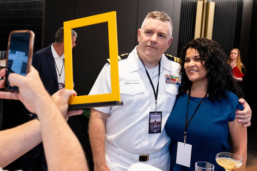 A man in a formal military uniform poses for a photo with a woman in business attire as another person, not pictured, holds up a phone and a yellow, square picture frame.