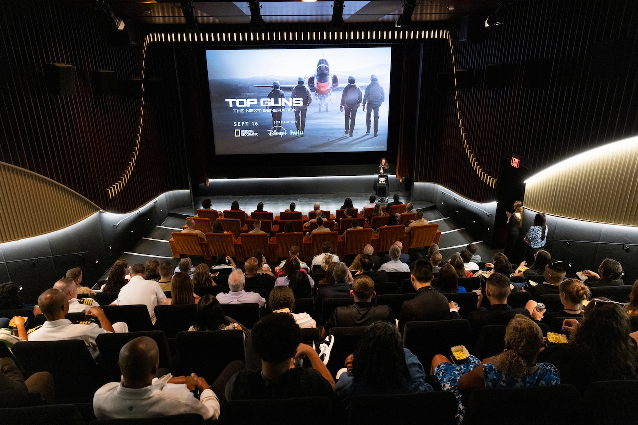 A person stands at a podium at the front of a movie theater filled with people. The screen reads, “Top Guns: The Next Generation” and depicts four people walking toward an aircraft.