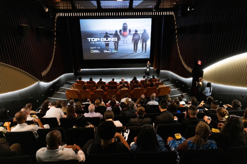A person stands at a podium at the front of a movie theater filled with people. The screen reads, “Top Guns: The Next Generation” and depicts four people walking toward an aircraft.