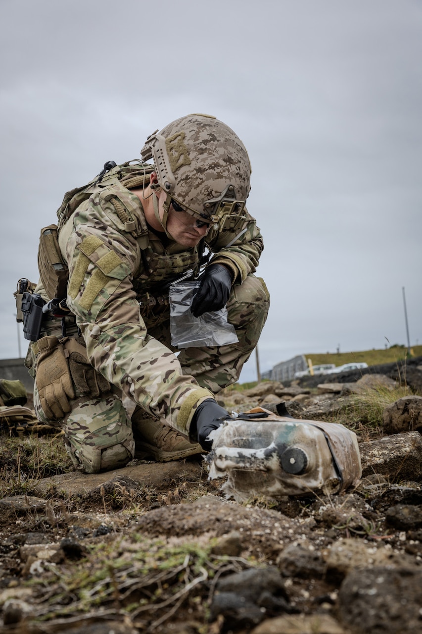 A person in a bomb suit kneels down to pick up a bag lying on the ground.