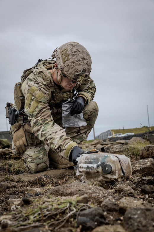 A person in a bomb suit kneels down to pick up a bag lying on the ground.
