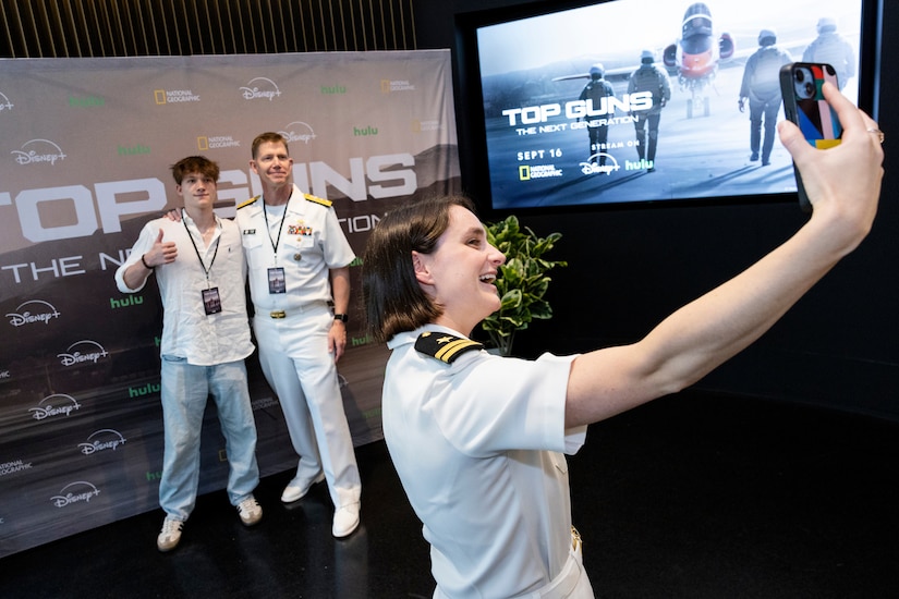 A woman wearing a formal military uniform takes a selfie with two men in the background, standing in front of a large poster. One of the men is dressed in casual attire, and the other is wearing a formal military uniform.