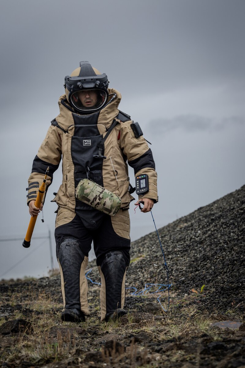 A person in a bomb suit walks across a field. The person is holding a tool.