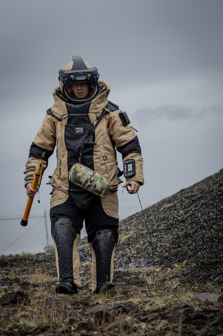 A person in a bomb suit walks across a field. The person is holding a tool.