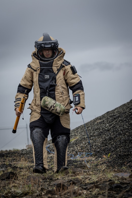 A person in a bomb suit walks across a field. The person is holding a tool.