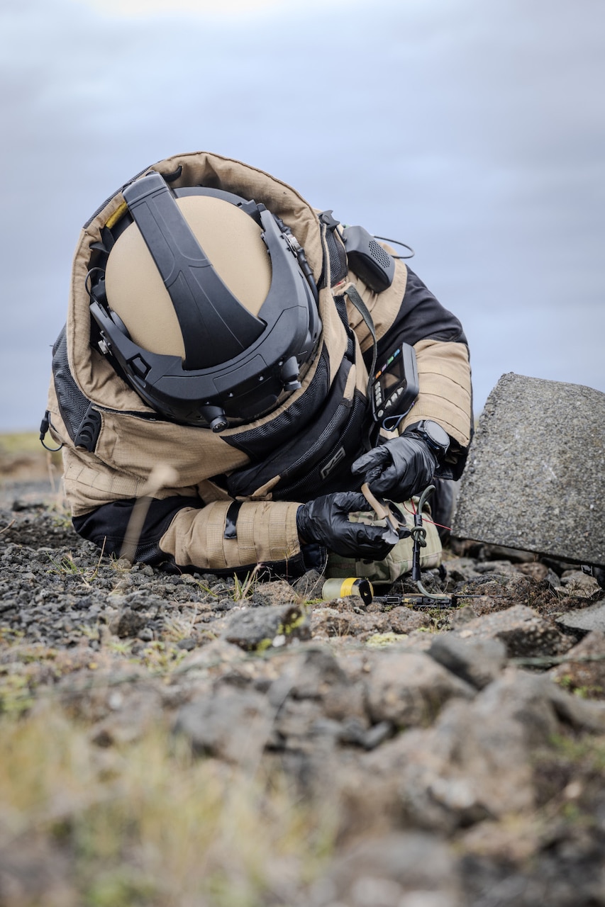 A person in a bomb suit kneels down to pick up a bag lying on the ground.