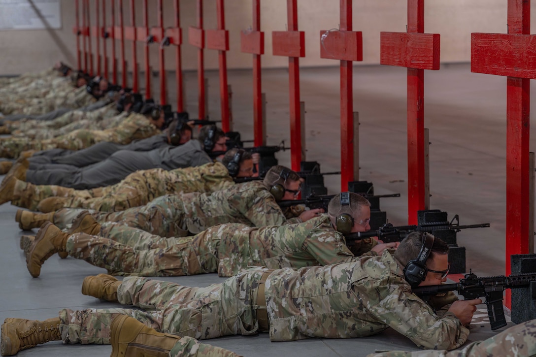 A large group of airmen lies on concrete and aims service weapons on red wooden structures.