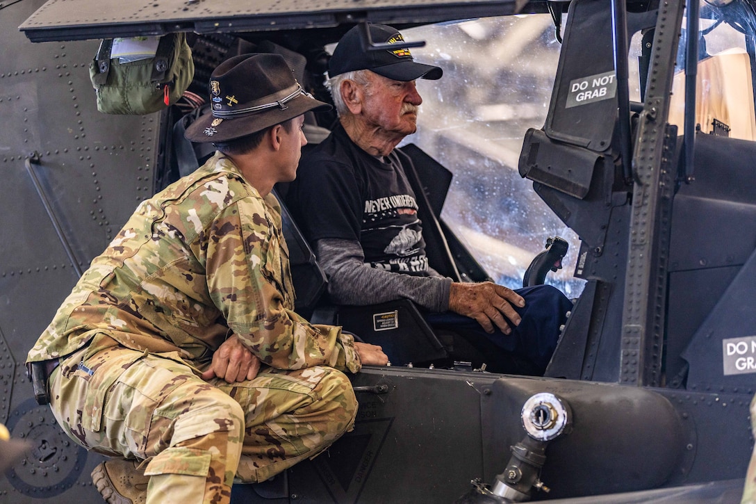 A person wearing dark clothes and a baseball cap sits in a parked aircraft while a soldier leans over.