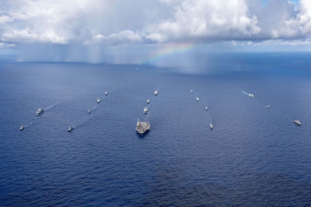 A large group of ships steams in five columns in front of rain clouds and a rainbow on the ocean.