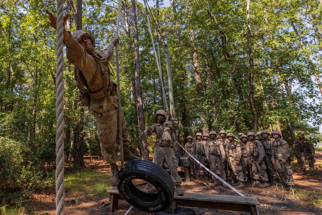 A Marine Corps recruit reaches toward a rope while holding on to another rope on an obstacle course while a group of recruits watches from behind in a forest setting.