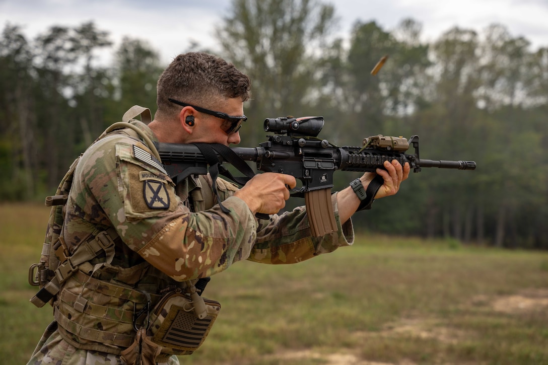 A soldier aims and shoots a service weapon outside in a field with trees in the background during the day.