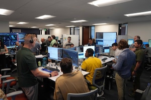 A group of people in business casual attire stand and sit in a room in front of computer screens.
