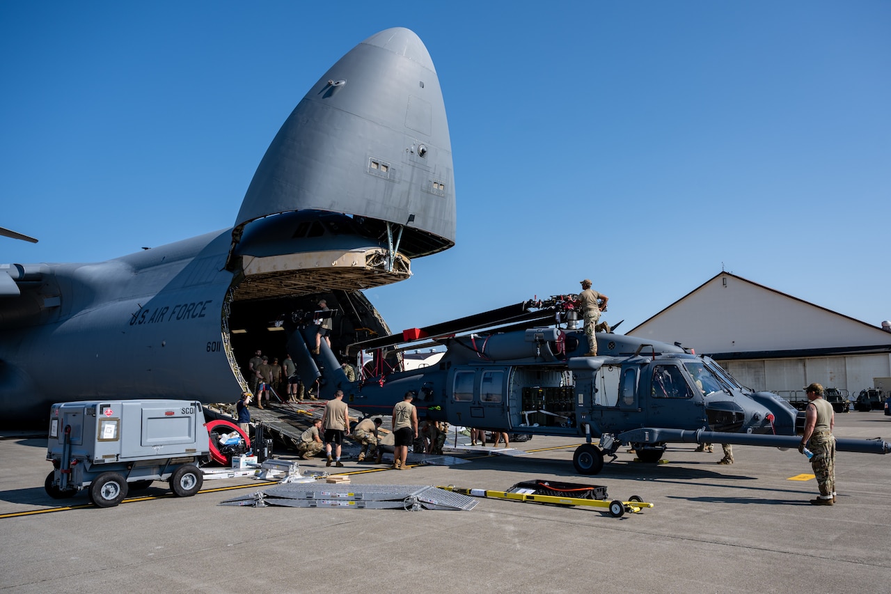 A handful of service members in casual attire are on a tarmac loading a military helicopter with its rotors folded together into the front end of a large military cargo aircraft.