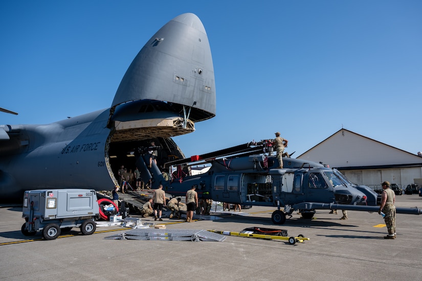 A handful of service members in casual attire are on a tarmac loading a military helicopter with its rotors folded together into the front end of a large military cargo aircraft.