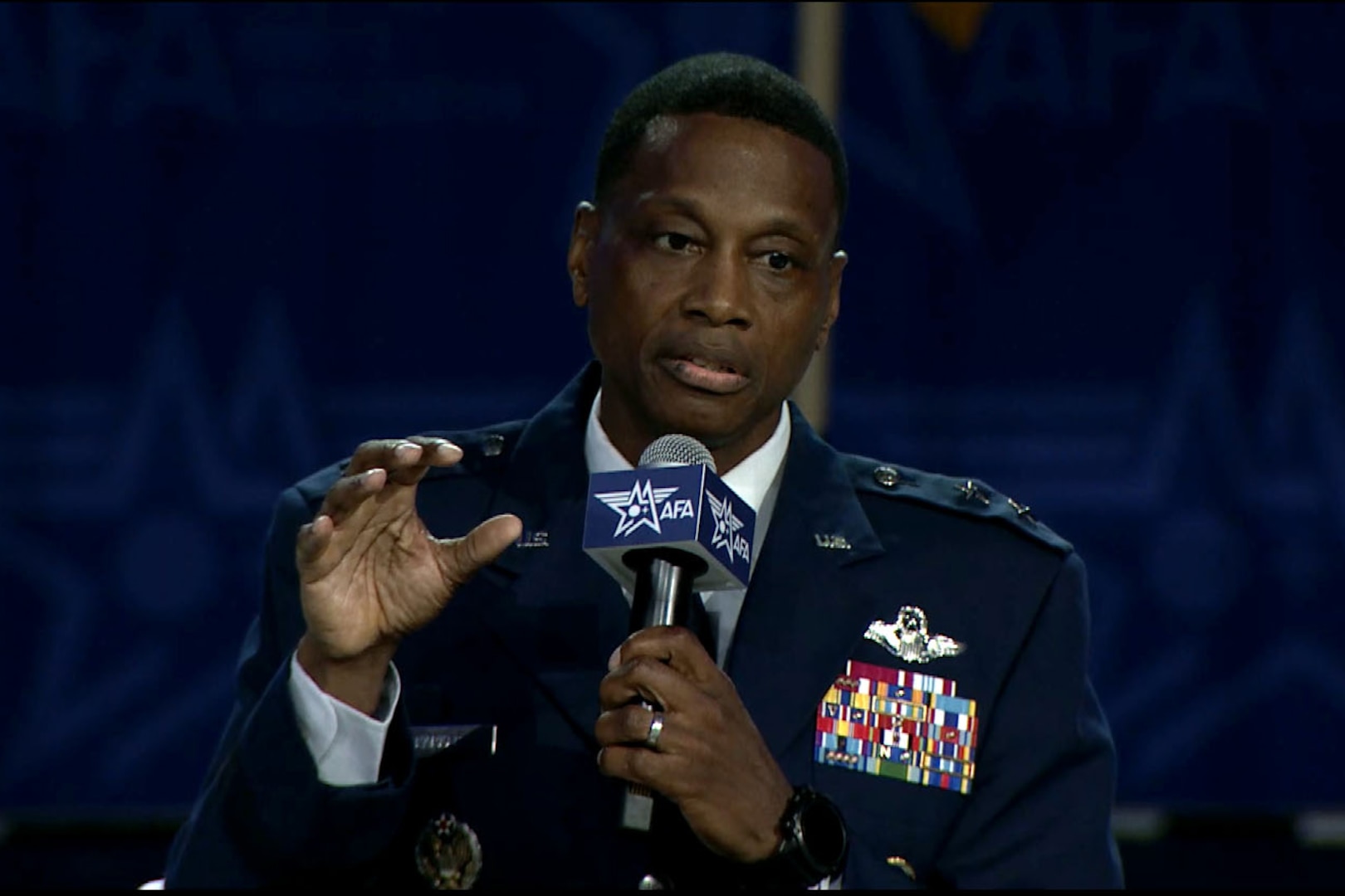 A man seated in a dress uniform speaks into a microphone and gestures.