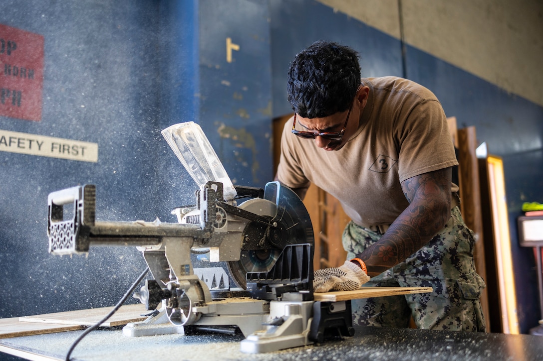 U.S. Navy Petty Officer 2nd Class Juan Delarosa, a utilitiesman with Assault Craft Unit 5, Naval Beach Group 1, saws plywood to build portable air conditioning unit stands during Operation Clean Sweep III at Marine Corps Base Camp Pendleton, California, Sept. 19, 2025. OCS III, part of the ongoing Barracks 360 Reset initiative, demonstrates a collective commitment to improving living conditions through shared ownership, sustained accountability, and tangible improvements to the barracks. Delarosa is a native of Connecticut. (U.S. Marine Corps photo by Sgt. Bryant Rodriguez)