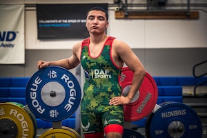 A man in a wrestling singlet leans against a rack of weights inside a gym