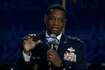 A man seated in a dress uniform speaks into a microphone and gestures.