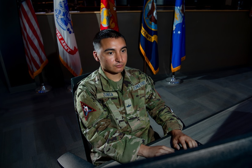 A man wearing a camouflage military uniform types while looking at a computer screen. There are military flags displayed in in the background.
