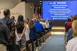 Back view of people standing in auditorium pacing stage with large blue screen with white words