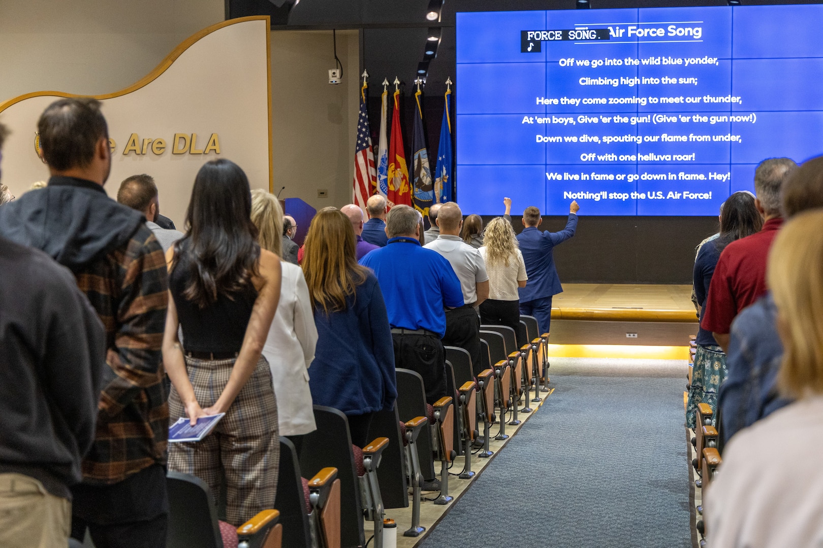 Back view of people standing in auditorium pacing stage with large blue screen with white words
