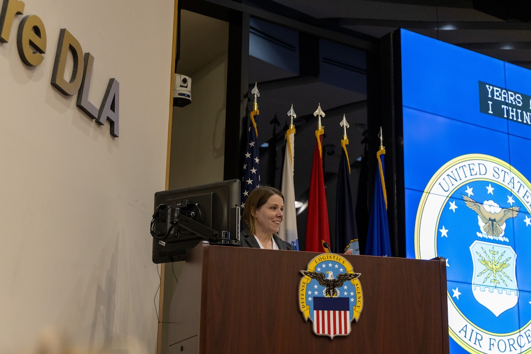 Close up of woman smiling behind podium with DLA emblem on it. Flags in background to the right.