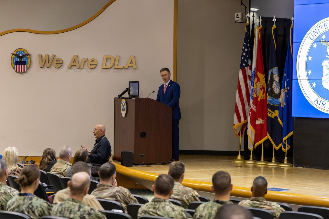 Man in blue suit stands behind podium on stage with flags to the right and 'We are DLA' on the wall to the left