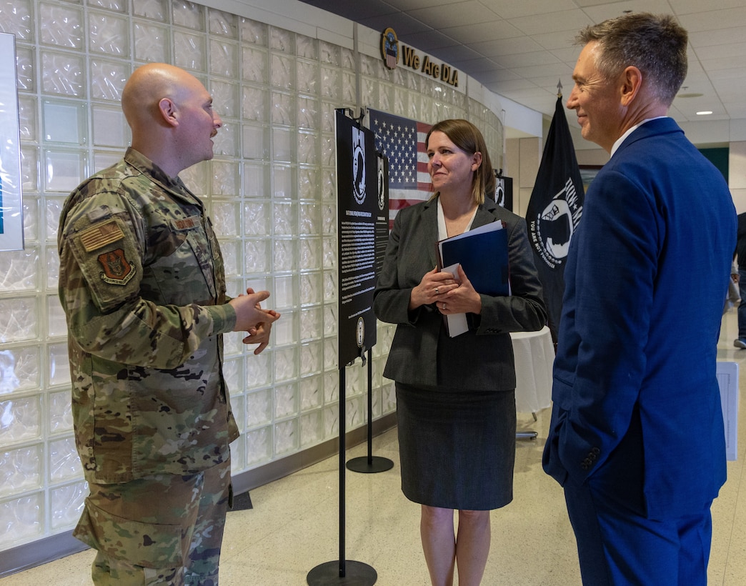 Man in military uniform to the left, woman in black skirt suit in the center and man in blue suit on the right, all in conversation.
