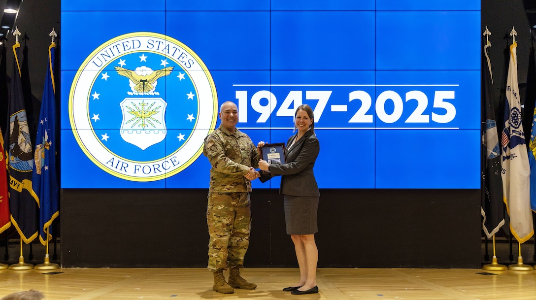 Man in military uniform on left and woman wearing black skirt and jacket on right, standing on stage shaking hands and holding framed certificate. Blue screen behind with Air Force emblem