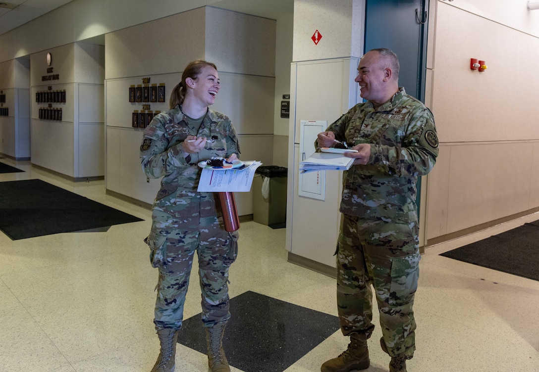 Woman on left, man on right, both wearing military uniform, looking toward each other smiling and holding cake and papers.