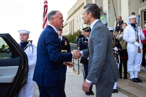 Two men dressed in business suits shake hands in front of a building. There is a sailor wearing a military dress uniform holding a car door to the left and several other service members in dress uniforms standing behind the two men.