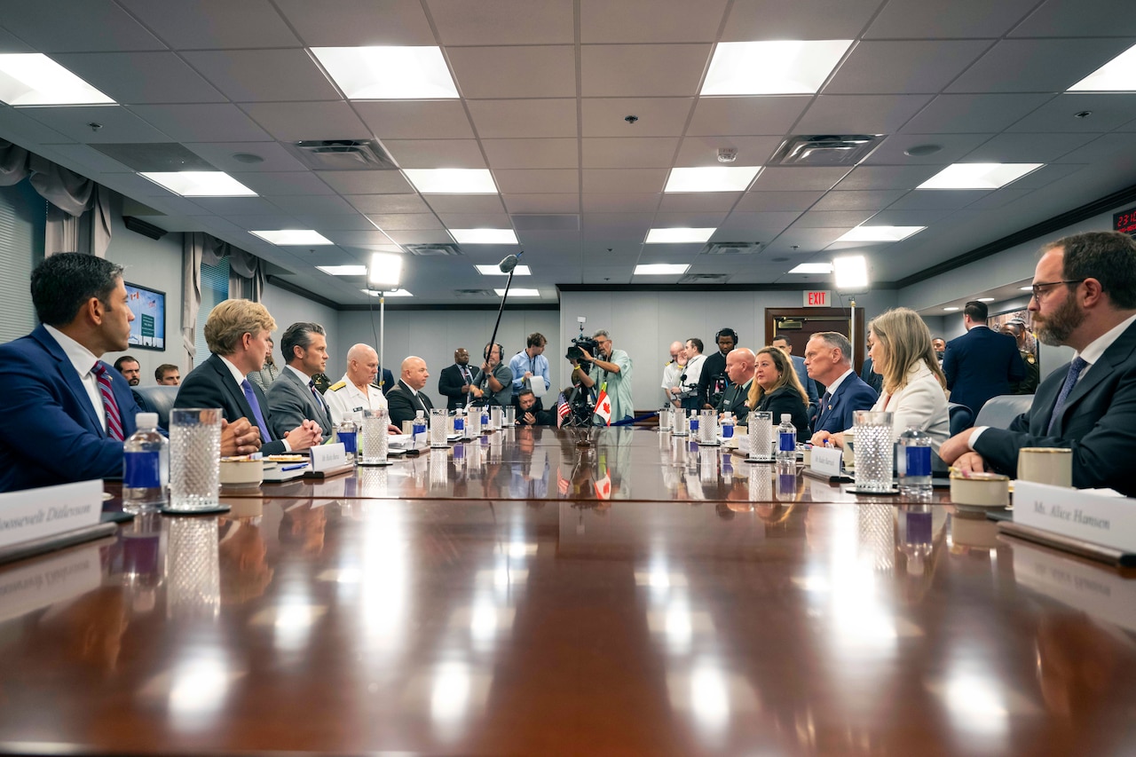 Men and women in business attire sit around a long wooden table. There are three people in business casual attire at the end of the table holding cameras and one person holding a shotgun microphone.