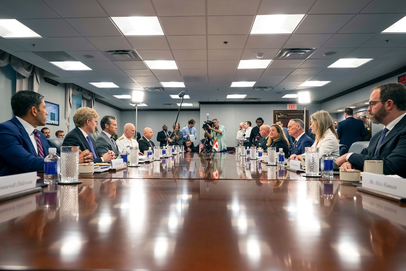 Men and women in business attire sit around a long wooden table. There are three people in business casual attire at the end of the table holding cameras and one person holding a shotgun microphone.