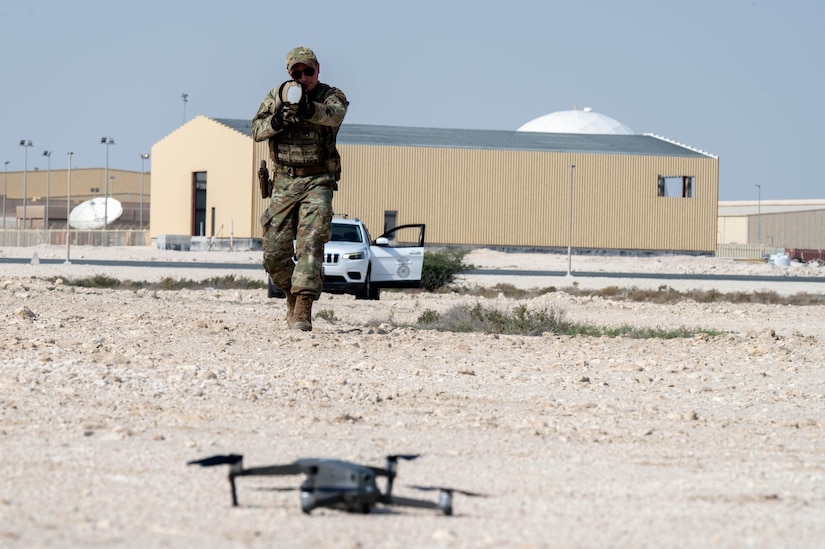 A person wearing a camouflage military uniform points a weapon at a small drone sitting on the ground in a desert environment.