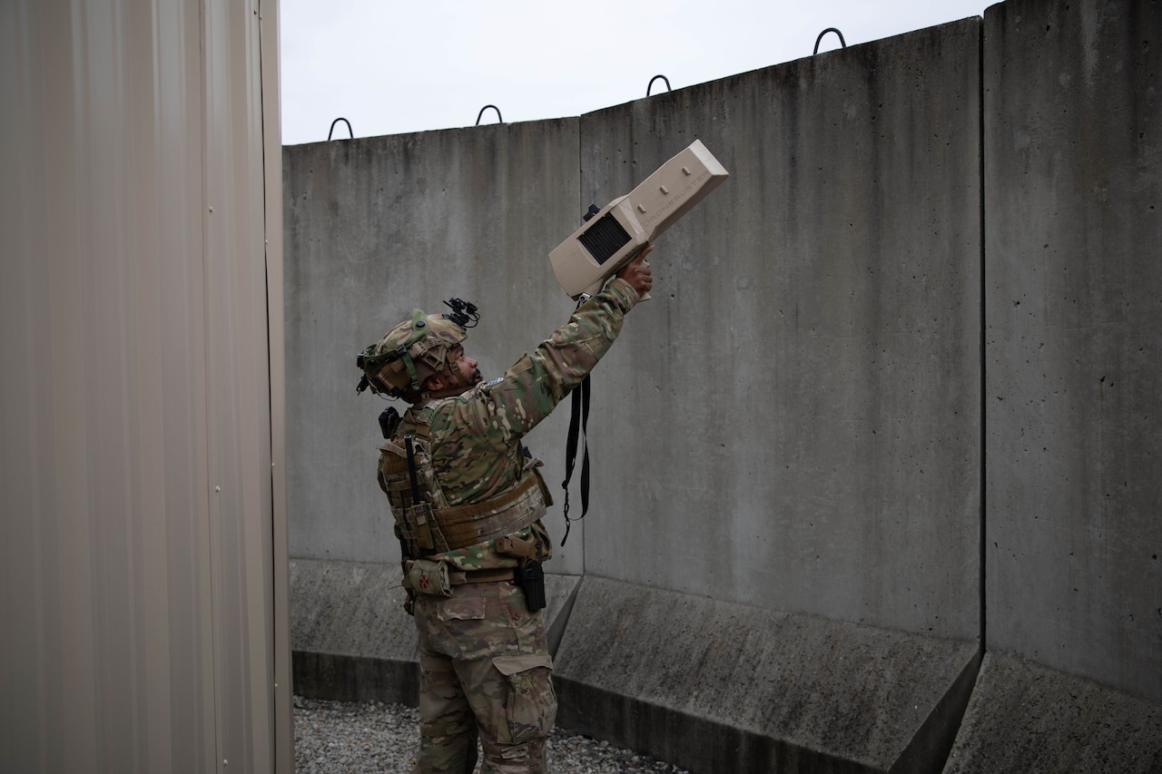 A man in a camouflage military uniform points a handheld rectangular weapon device above a concrete wall.