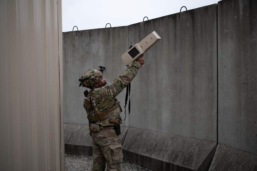 A man in a camouflage military uniform points a handheld rectangular weapon device above a concrete wall.